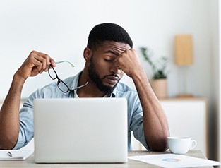 Tired man sitting at laptop rubbing eyes while holding glasses